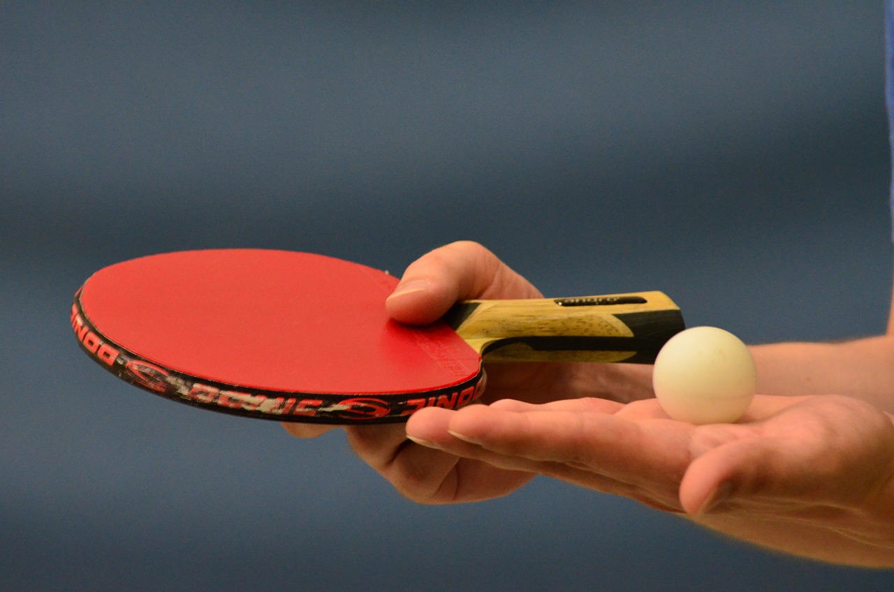 Tischtennisschläger mit rotem Belag in der linken Hand eines Mannes, in der rechten Hand ein cremefarbener Tischtennisball, beides flach gehalten, um den Ball anzuschneiden; zu sehen sind nur die Hände, der Hintergrund ist verschwommen.